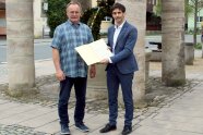 Zwei Männer stehen vor dem Brunnen am zentralen Platz in der Ortschaft Baudenbach und freuen sich mit einem Zuwendungsbescheid für ein Pressefoto über EU-Fördermittel.