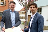 Zwei Männer stehen in Dietersheim im Herzen der Ortschaft vor dem festlich geschmückten Osterbrunnen und freuen sich mit einem Zuwendungsbescheid für ein Pressefoto über EU-Fördermittel.