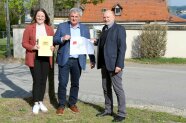 Drei Menschen haben vor einem Friedhof in Arberg einen Zuwendungsbescheid für ein Pressefoto in der Hand und freuen sich über EU-Fördermittel.