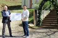 Zwei Männer stehen in Bergen vor der alten, schmalen Treppe, die zur Kirche des Dorfs führt und freuen sich mit einem Zuwendungsbescheid für ein Pressefoto über EU-Fördermittel.