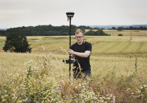 Ein Mann steht in einem Feld aus Blunen und Roggen. Er hält ein GPS-Gerät in der Hand.