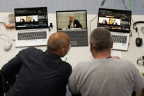 Zwei Männer sitzen vor drei Laptops, auf denen die hybride Veranstaltung übertragen wird.