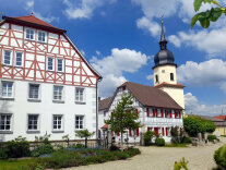 Der Dorfplatz in Auernhofen ist mit Sitzmöglichkeiten und Brunnen der Mittelpunkt der kleinen Ortschaft. Darum herum stehen viele Gebäude mit schmuck sanierten Fassaden.