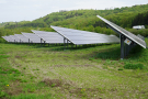 Freiflächen-Photovoltaikanlage im Vordergrund. Im Hintergrund bewaldeter Hügel. Weißer Laster fährt auf der nahen Autobahn.