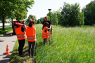 Eine Schülerin steht am GPS-Gerät und misst, während zwei weitere mit Pflöcken bewaffnet auf das Ergebnis warten.