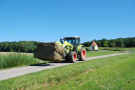 Ein Traktor, auf dessen Frontlader zwei Heuballen übereinander geladen sind, fährt auf einem Asphaltweg. Ein kleiner Stichweg führt zu einer Kapelle.