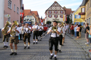 Eine Musikkapelle zieht in einem Festumzug durch die Straßen einer fränkischen Ortschaft. Das Publikum verfolgt das Treiben vom Straßenrand aus.